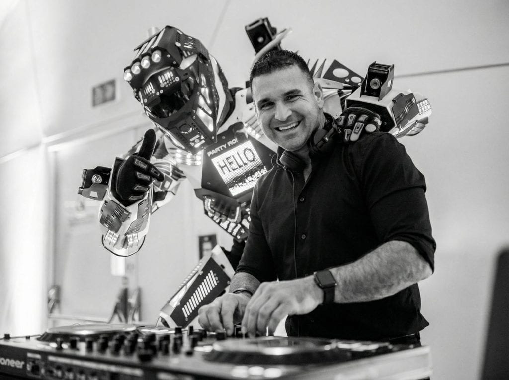 Félszemű DJ és egy robot táncolnak, miközben zenét játszanak, jó hangulatban.