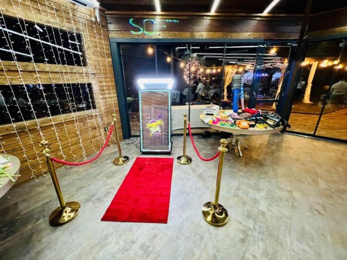Modern fotósarok LED-es tükörrel, vörös szőnyeggel, arany oszlopokkal és party kellékekkel asztalon.