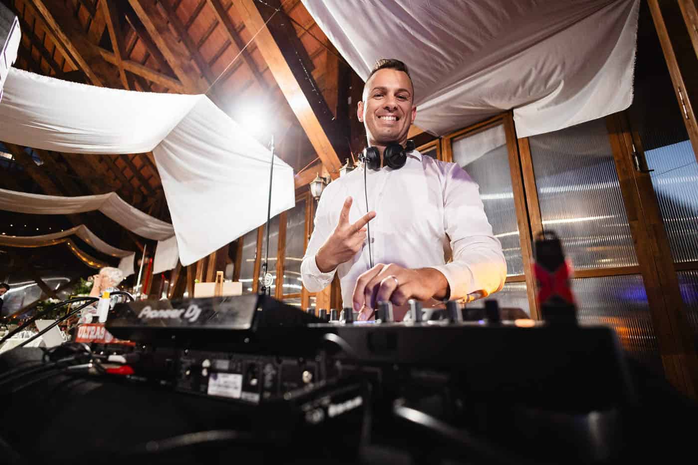 DJ keverőpultnál áll egy fa szerkezetű, lakkozott tetős helyiségben, békejelet mutatva.