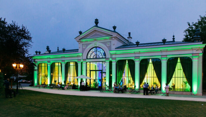 Tata Pálma Rendezvényház - Angolkert - Imposing building with green and yellow lights, large windows, people gathered outside.
